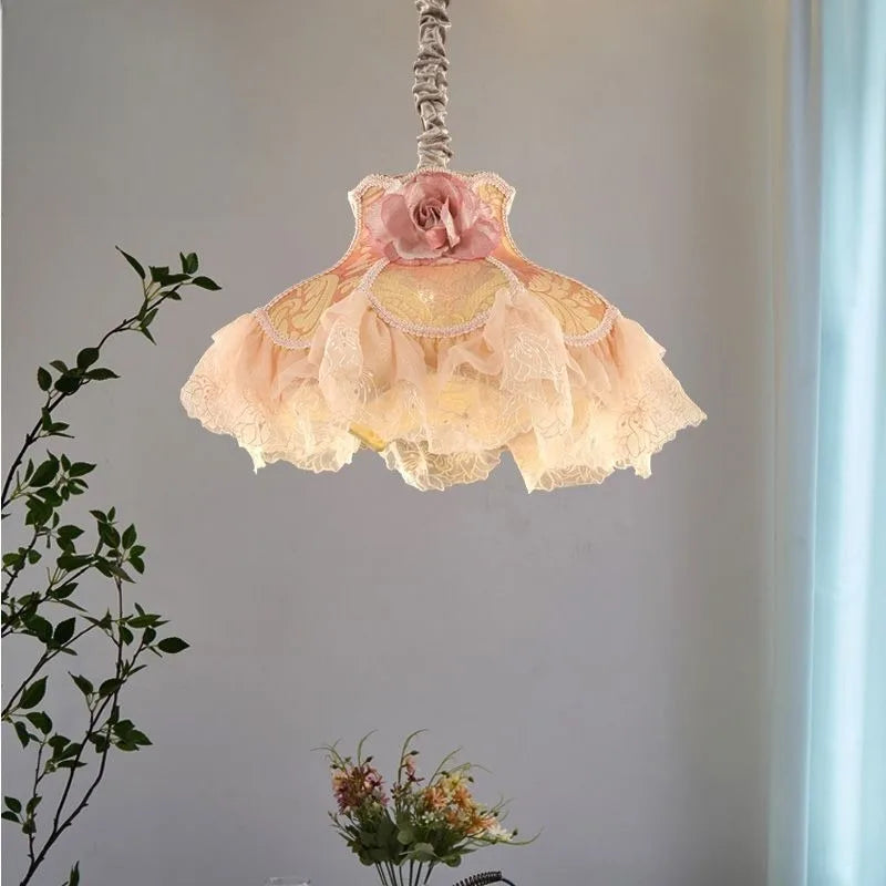A fabric pendant light with a pink rose accent and ruffled shades hangs in a softly lit room with greenery in the foreground.