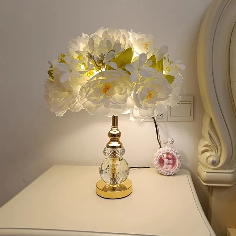 A bedside table with a white floral table lamp with a crystal base, casting a bright light.