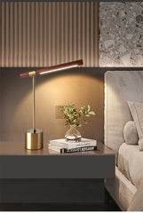 Brass and wood LED desk lamp with the light on, situated on a nightstand next to a bed and books.