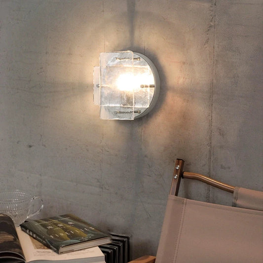 A modern silver wall sconce with clear glass panels mounted on a grey textured wall above a chair and books.
