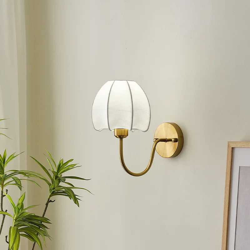 Close-up of a gold wall sconce with a white pleated fabric shade shaped like a flower, showing the mounting plate and curved arm.