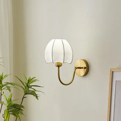 Close-up of a gold wall sconce with a white pleated fabric shade shaped like a flower, showing the mounting plate and curved arm.