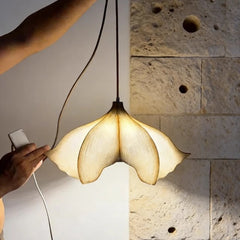 A hand holding a remote to a beige floral pendant light, illuminated against a textured stone wall.