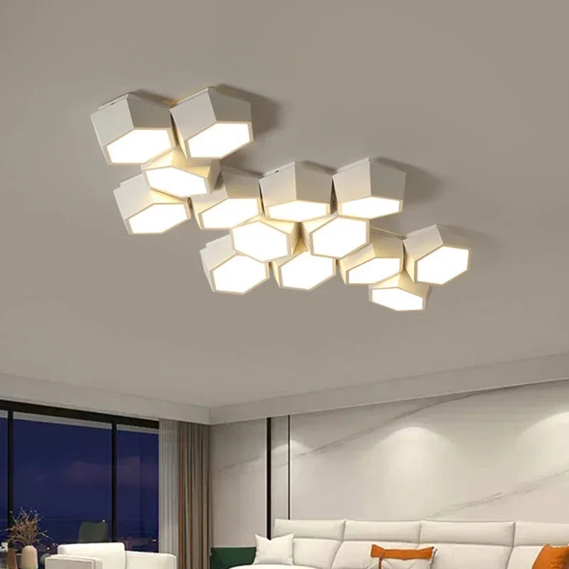 A modern white geometric cluster ceiling light with hexagonal panels emitting a soft white light, mounted on a white ceiling in a contemporary living room with a sofa visible.
