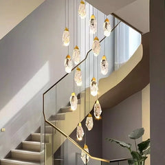 A modern interior staircase with glass railings, illuminated by a tall, cascading gold chandelier with faceted crystal pendants.