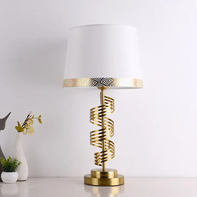 A gold spiral table lamp with a white conical fabric shade sits on a surface against a white wall.