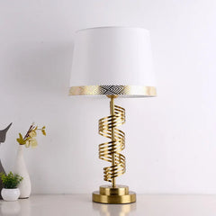 A gold spiral table lamp with a white conical fabric shade sits on a surface against a white wall.
