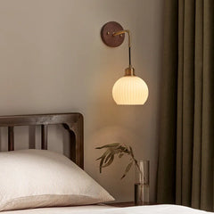 A bedroom scene with a walnut wood wall sconce, its frosted glass shade casting a warm glow near a bed and a vase of greenery.