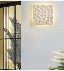 A light stone textured wall sconce mounted on a white wall next to a large window with palm leaves visible inside.