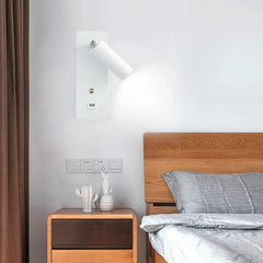 A white wall sconce with a white adjustable spotlight head and integrated USB port and switch, mounted next to a wooden bed.