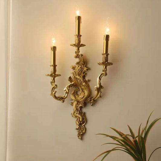 Illuminated gold candelabra wall sconce with three candle-shaped lights, casting a warm glow on a wall, with a plant in the foreground.