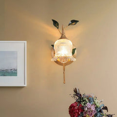 Gold flower wall sconce with clear glass shade on a beige wall, near a framed picture and a bouquet of flowers.