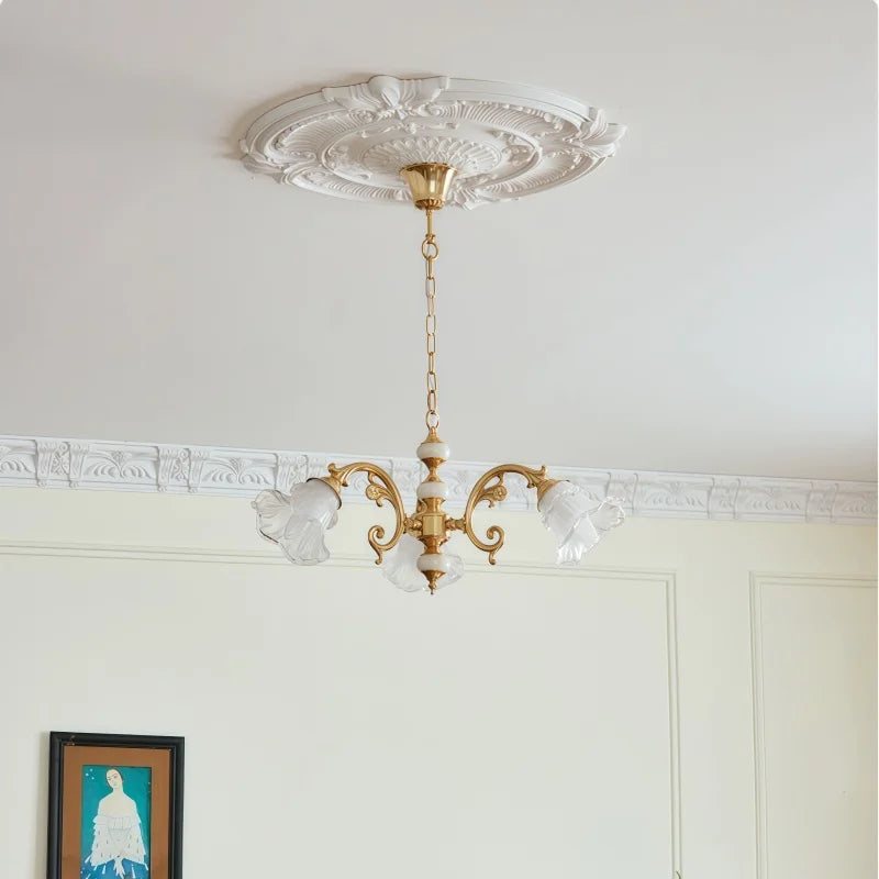 A gold chandelier with three frosted glass flower-shaped shades, suspended from a white ceiling with decorative molding.