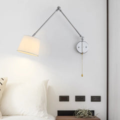Chrome swing arm wall sconce with a white fabric drum shade over a bedside table with electrical outlets.