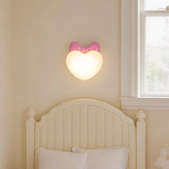 A pink bow heart-shaped wall sconce mounted on a cream-colored wall above a white headboard.