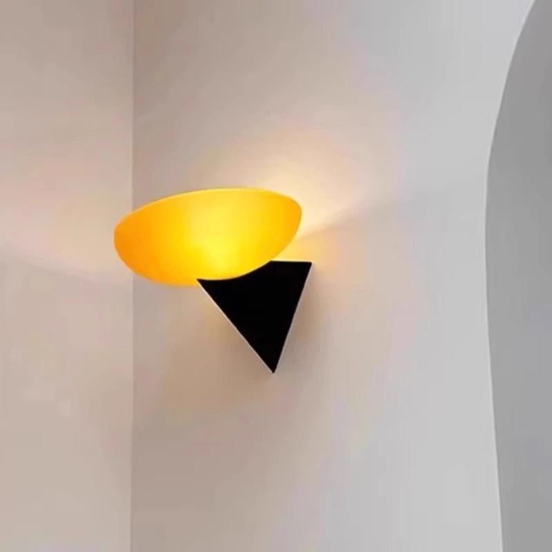 A contemporary wall sconce with a frosted yellow-toned oval shade and a black triangular base, illuminated in a corner of a room.