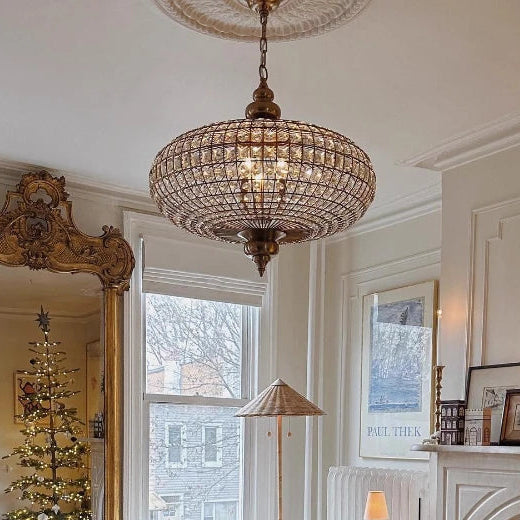 A large, ornate gold mirror reflects a Christmas tree and a living room setting, with a crystal orb chandelier hanging overhead.