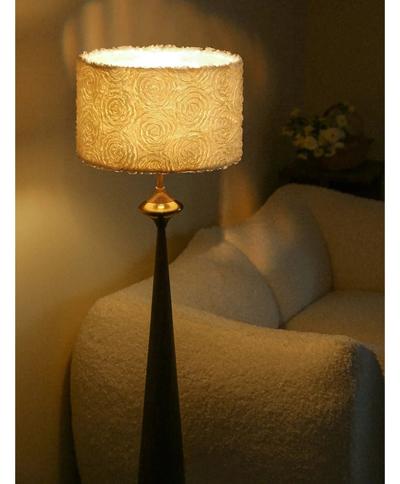 A floor lamp with a dark grey conical base and a drum shade covered in a textured floral pattern, illuminated from within.
