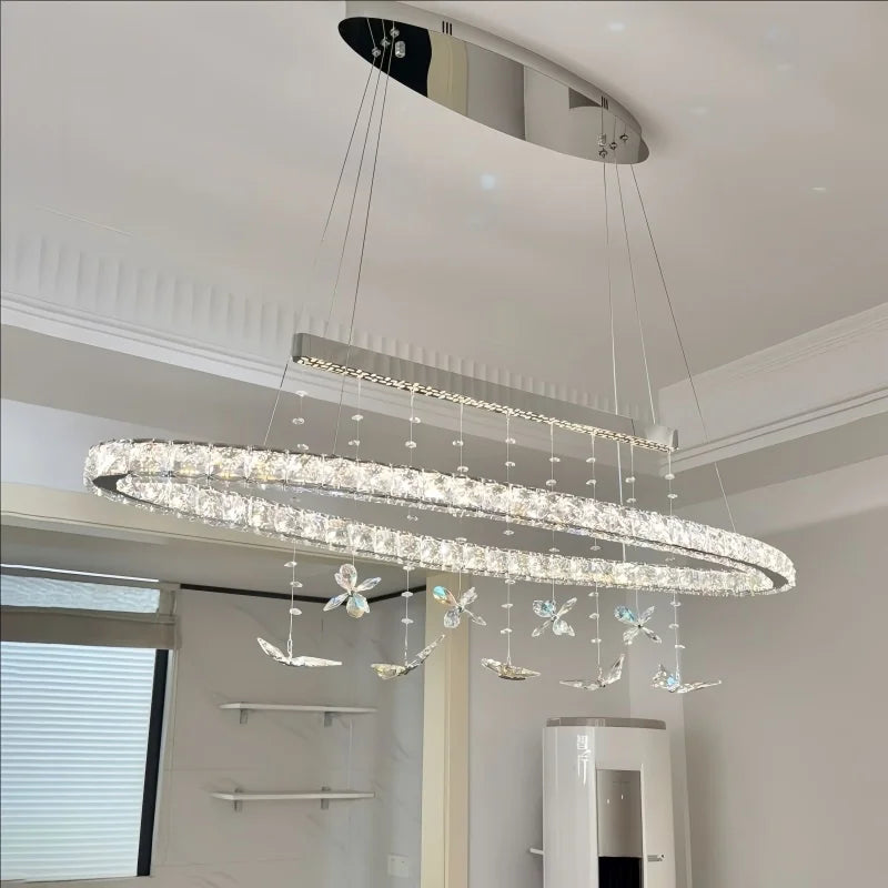 A long, oval-shaped crystal chandelier with hanging butterfly ornaments is shown illuminated in a modern interior setting.