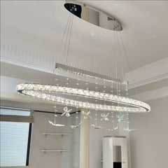 A long, oval-shaped crystal chandelier with hanging butterfly ornaments is shown illuminated in a modern interior setting.
