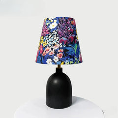 Black ceramic table lamp with a conical fabric shade displaying a vibrant, colorful floral pattern, placed on a white surface.