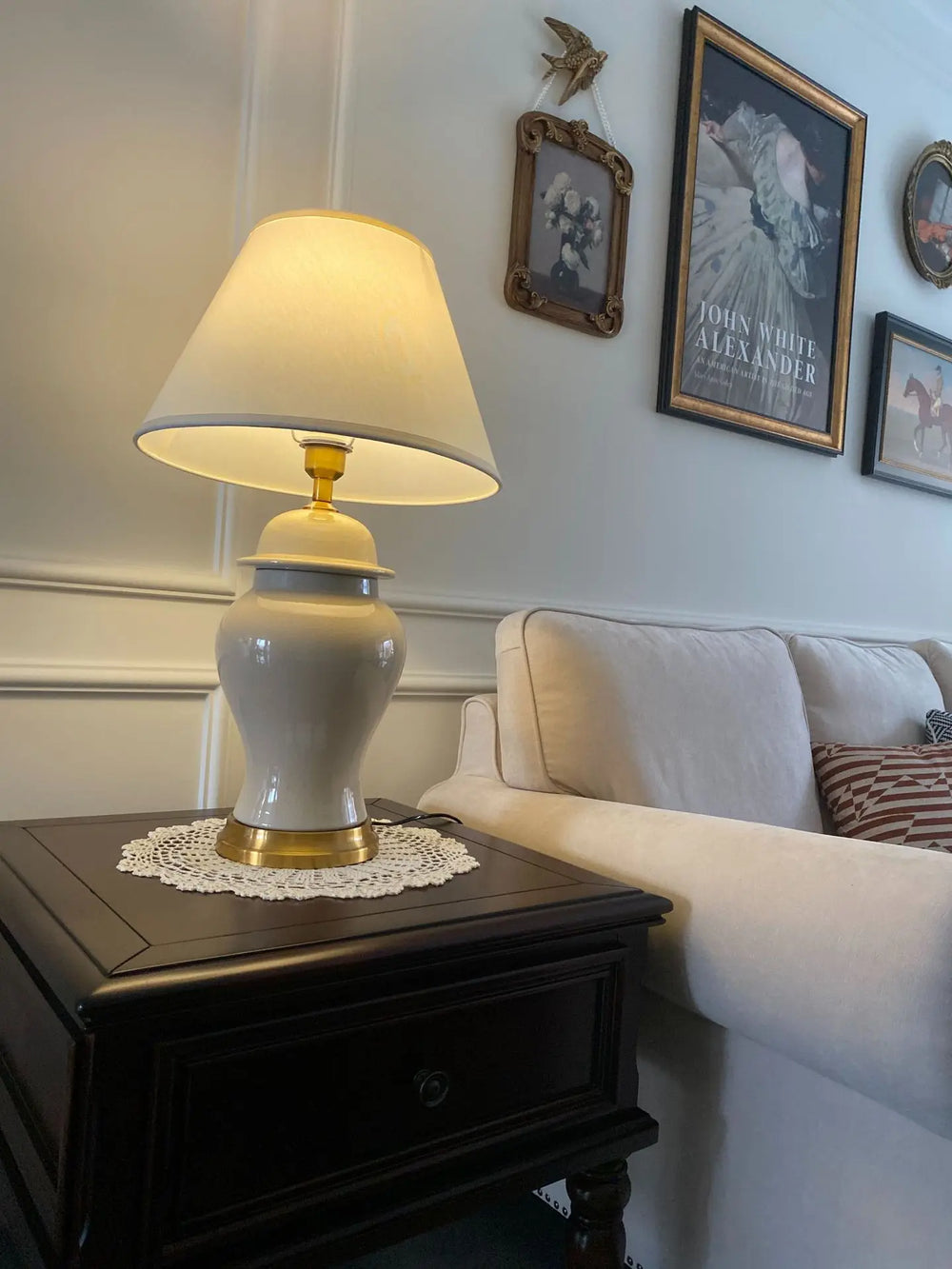 A white ceramic table lamp with a cream drum shade sits on a dark wooden side table next to a white sofa.