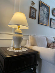 A white ceramic table lamp with a cream drum shade sits on a dark wooden side table next to a white sofa.