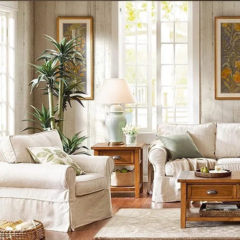 A light blue ceramic table lamp with a cream drum shade is placed on a wooden side table in a living room setting with white furniture and large windows.
