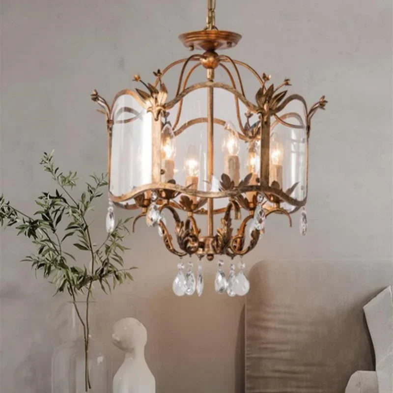 Gold candelabra pendant light with clear crystal droplets hanging from a distressed gold frame and a clear glass shade, illuminating a bedroom setting.