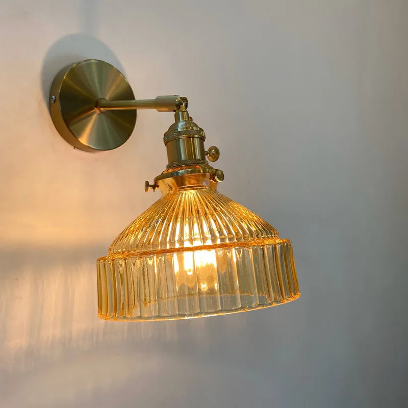 Amber fluted glass shade wall sconce with a brass arm and mounting plate illuminated against a beige wall.
