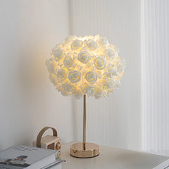 A white floral rose bud table lamp with a gold base and stem, illuminated on a white surface.