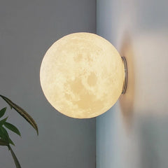 A 3D printed moon shaped wall sconce with a textured surface is mounted on a gray wall, emitting a soft yellow light. A green leaf plant is visible in the foreground.