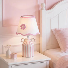 Pink and white striped ceramic table lamp with a scalloped white fabric shade featuring a pink flower accent, placed on a white nightstand next to a bed.