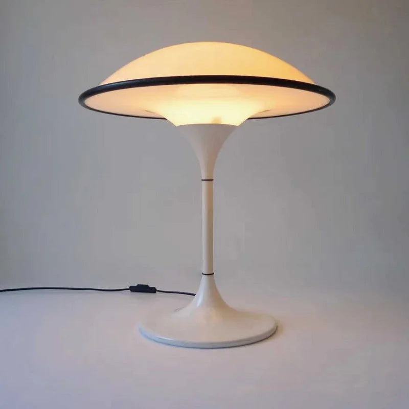 A lit white dome table lamp with a black rim, showing its base and stem, on a white surface with a power cord visible.