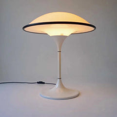 A lit white dome table lamp with a black rim, showing its base and stem, on a white surface with a power cord visible.