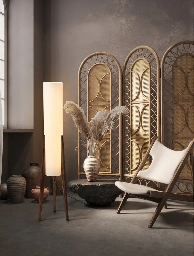 A tall floor lamp with a dark wood base and a white fabric shade, positioned in a dimly lit room with rattan screens and a canvas sling chair.