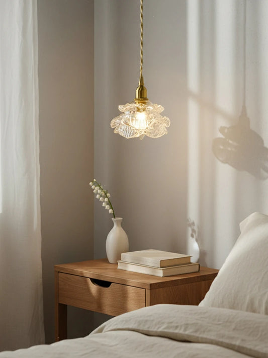 A brass pendant light with a crystal flower shade hanging above a bedside table with books and a vase.