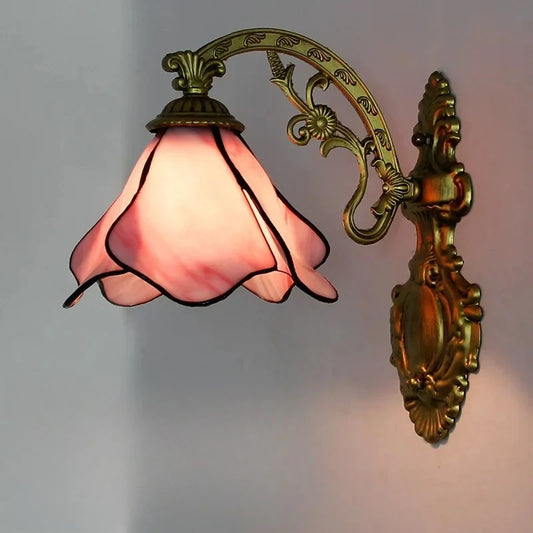 A wall-mounted light fixture with an antique gold metal arm and a scalloped pink stained glass shade, casting a soft glow against a light-colored wall.