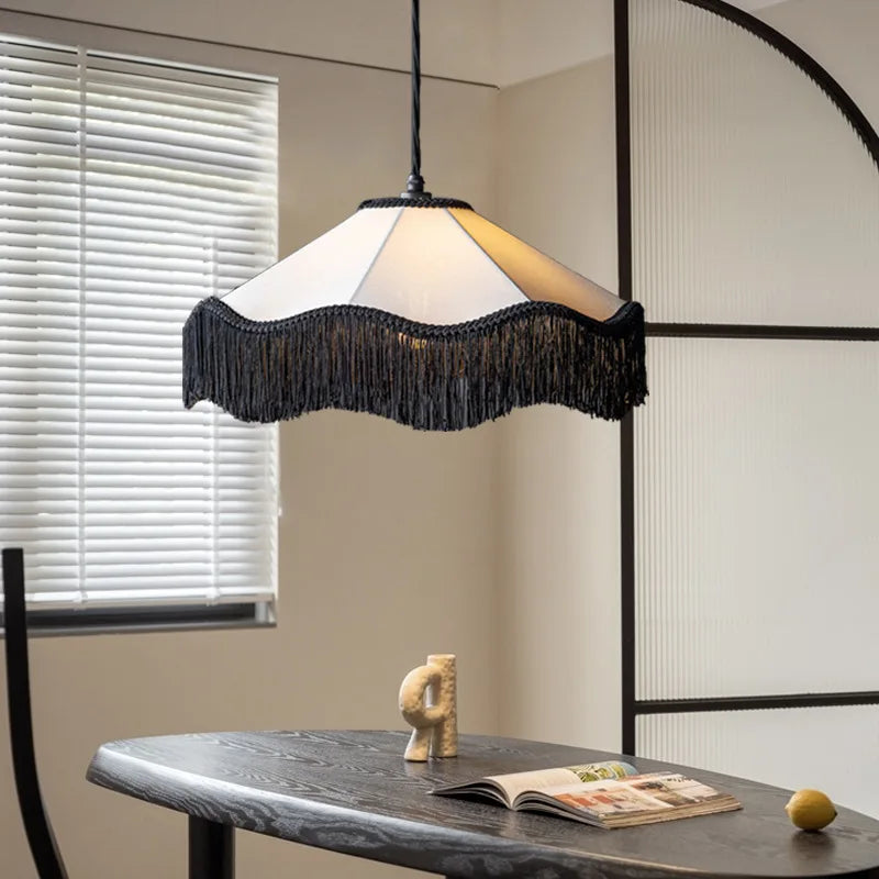 Fabric fringe pendant light with a white shade and black fringe, hanging above a dining table with blinds in the background.