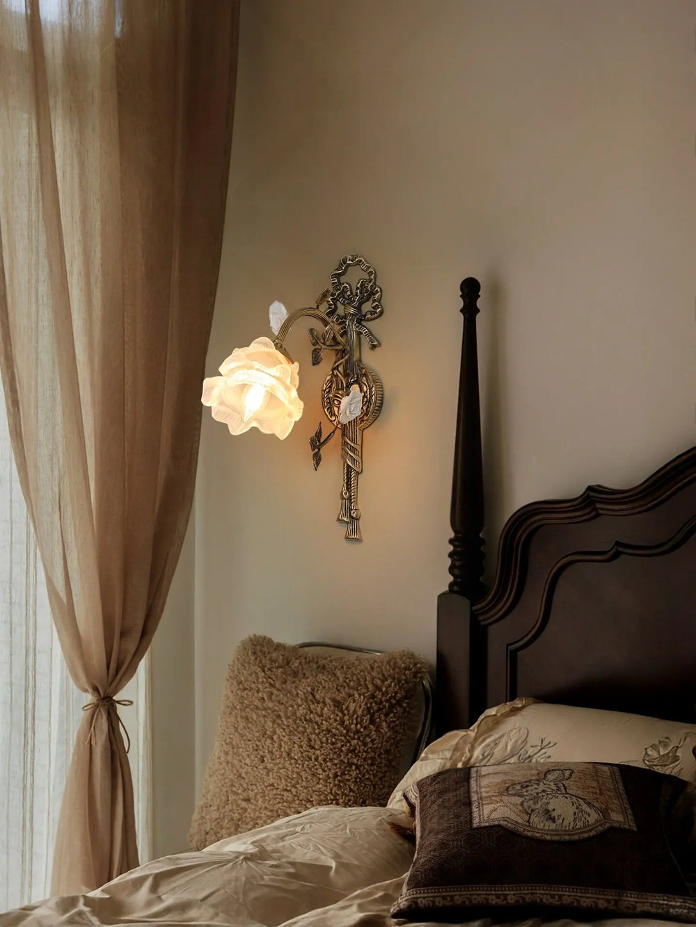 Antique gold flower wall sconce with a white frosted glass shade, illuminated in a bedroom setting with beige curtains and a detailed bed frame.
