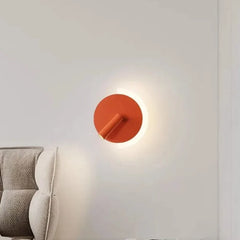 Orange modern LED wall lamp with a circular backplate and adjustable spotlight, mounted on a white wall next to a grey upholstered headboard.