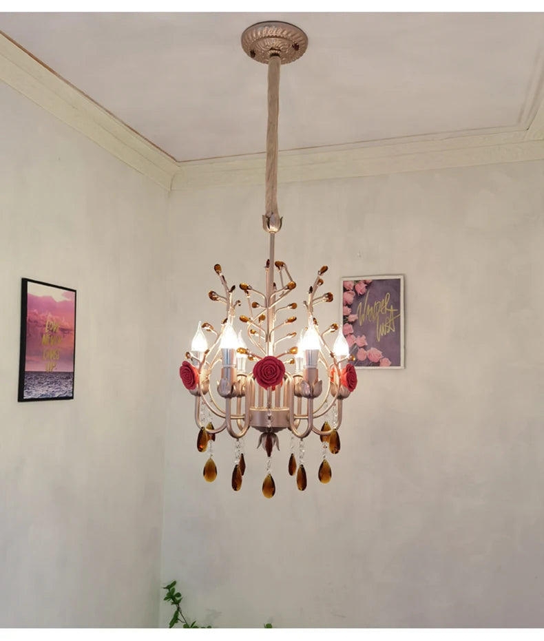 Pink and gold chandelier with ceramic roses and crystal droplets illuminated in a softly lit room with framed art on the wall.