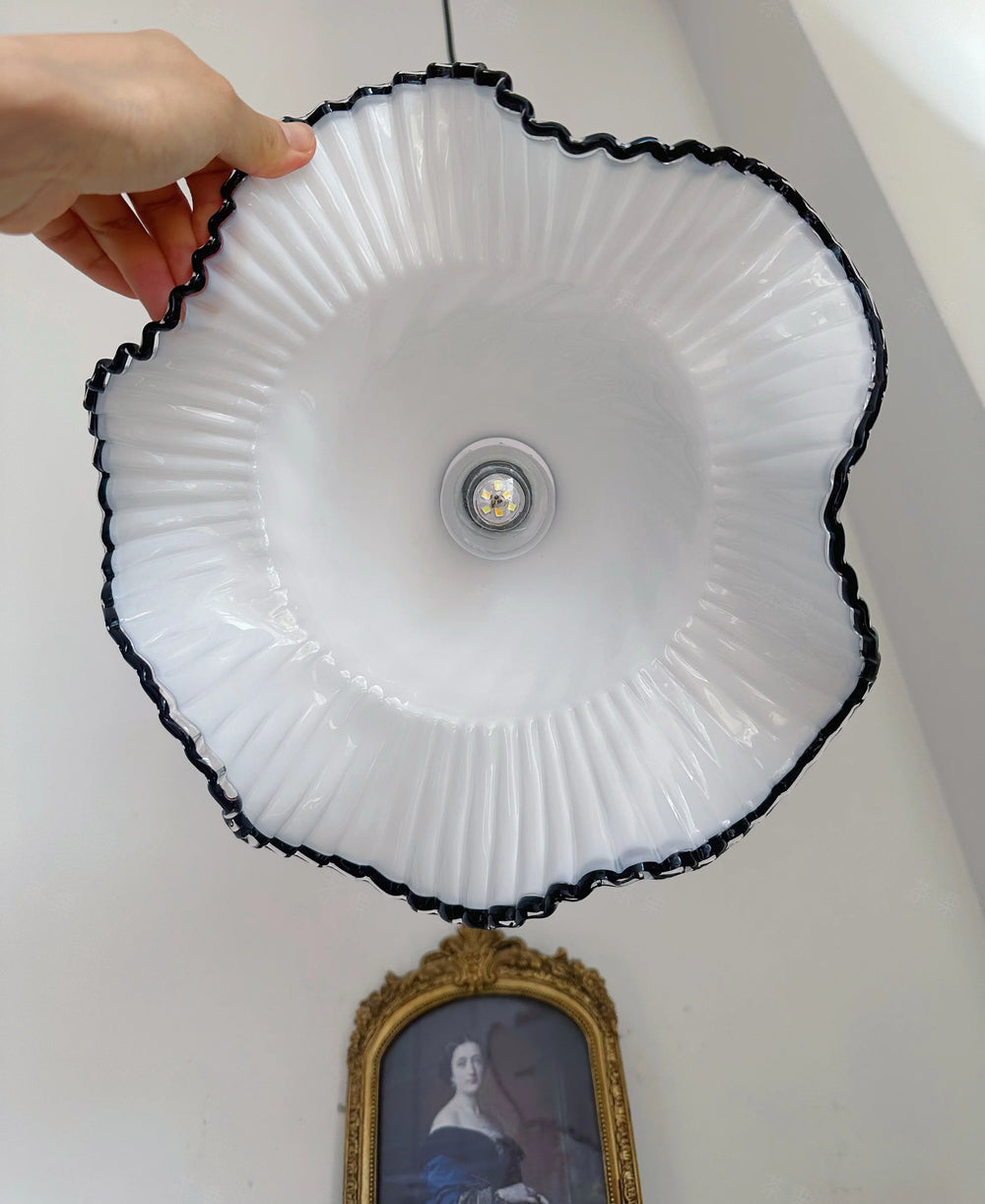 White light fixture with black rim held by a hand against a white wall with a portrait in the background.