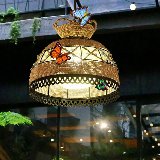A spherical, woven rattan pendant light with a decorative scalloped edge, featuring vibrant orange and green butterfly accents, suspended outdoors with a soft warm light.
