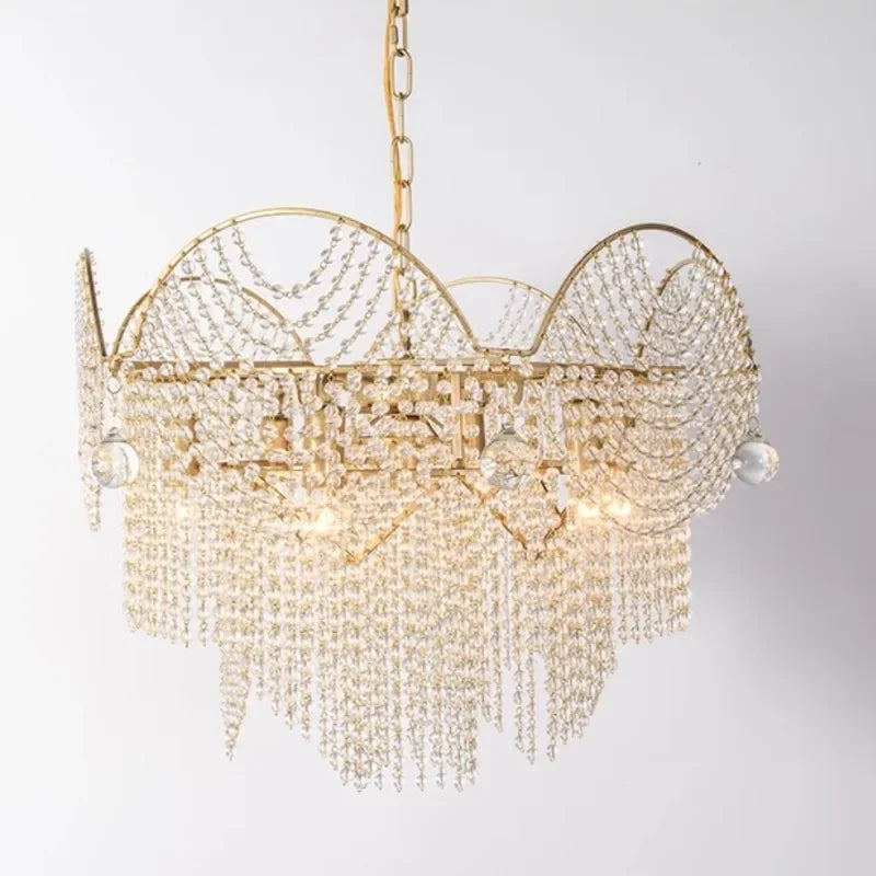 A close-up, front-facing view of a gold chandelier adorned with numerous clear crystals hanging in cascading strands and draped patterns against a white background.