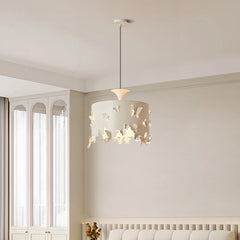 A cream-colored drum pendant light with butterfly cutouts, shown in a room with white curtains and furniture. The light illuminates the surrounding space.
