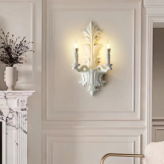 White leaf-shaped wall sconce with two candle-style lights mounted on a paneled wall, near a fireplace.