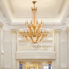 A grand gold tiered crystal candelabra chandelier hanging in a spacious hall with a balcony and marble columns.