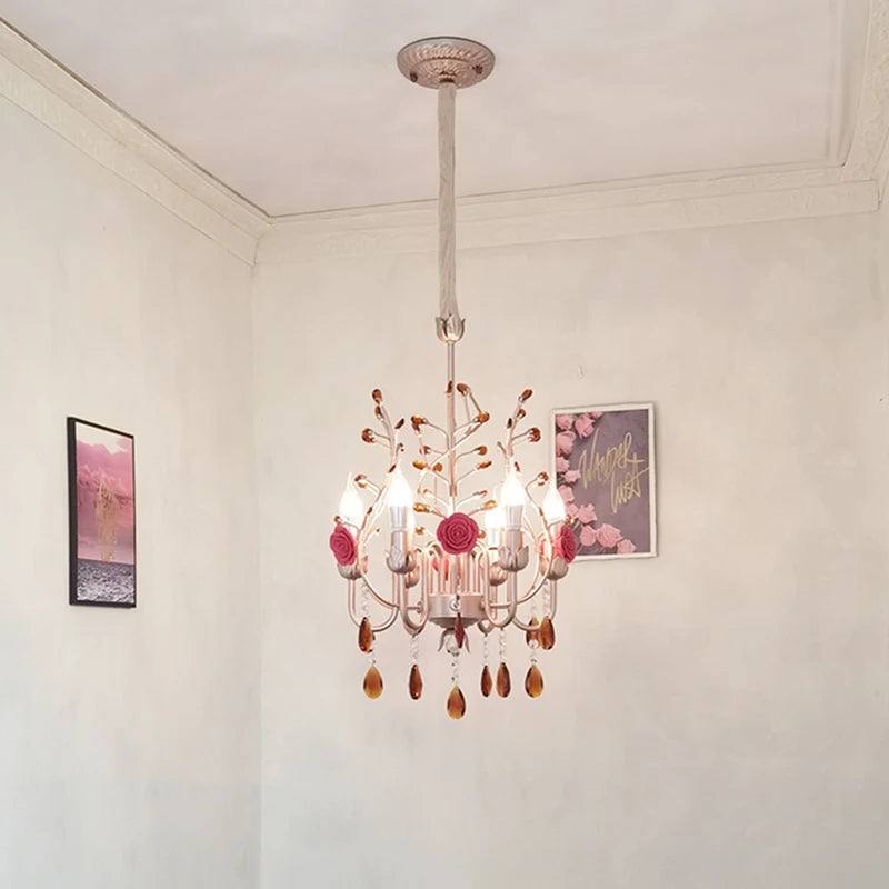 A pink and gold chandelier with rose and crystal details hangs from the ceiling in a room with white walls and decorative artwork.