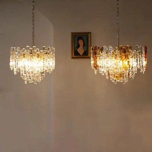 Two glass tube chandeliers with amber and clear glass tubes hanging on chains against a neutral wall with a framed portrait above.